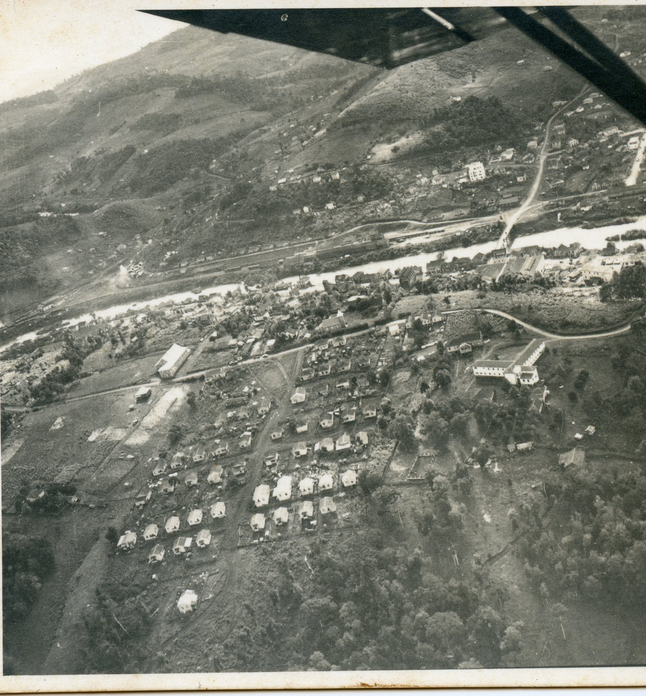 Fotografia aérea em preto e branco de uma pequena cidade brasileira, possivelmente em fase de expansão urbana inicial. A imagem revela um traçado urbano com casas dispostas de forma organizada, um rio atravessando a paisagem e áreas de vegetação nativa ao redor, sugerindo um contexto de colonização ou desenvolvimento regional do século XX.