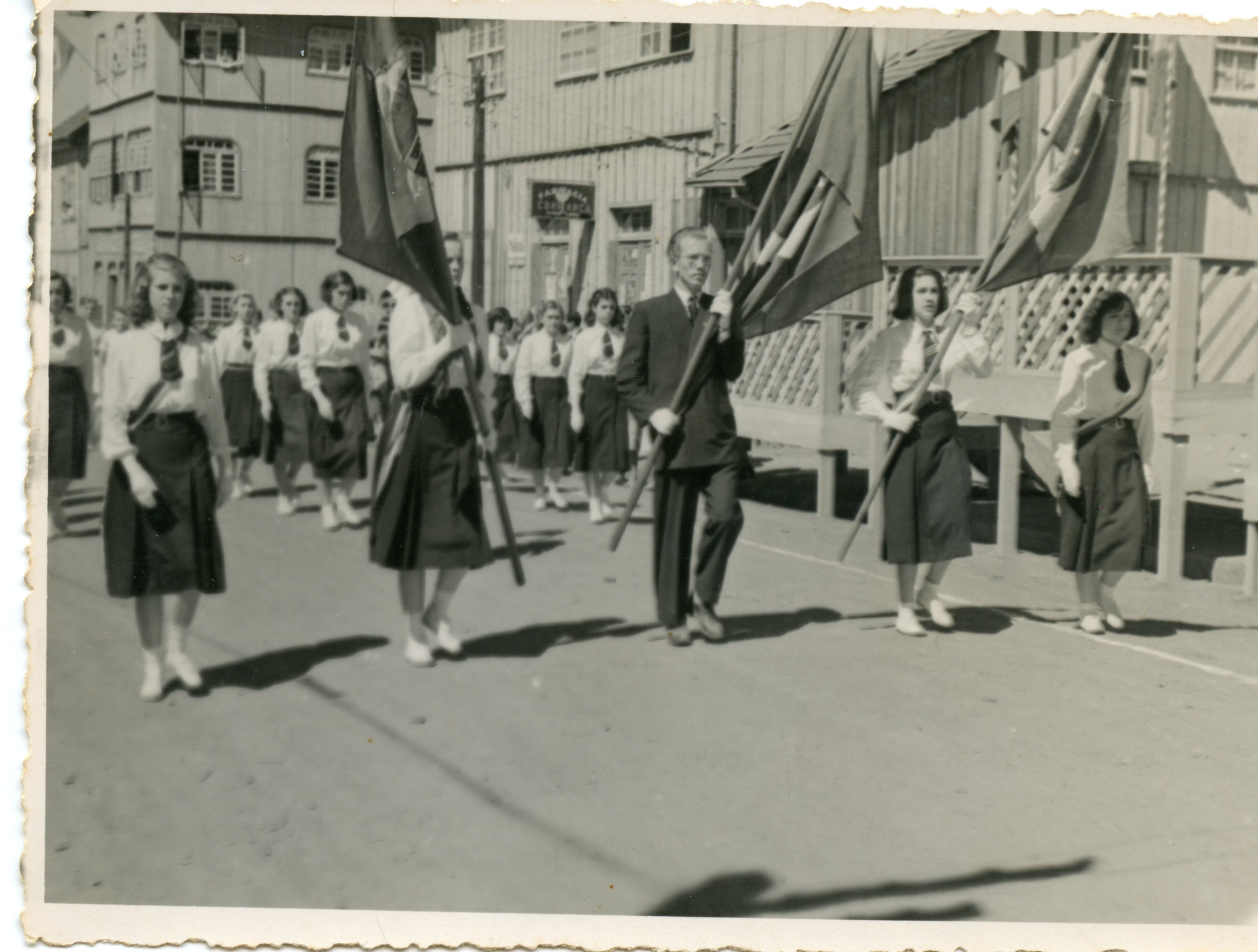Uma fotografia em preto e branco que retrata um desfile cívico ou escolar em uma rua com construções de madeira típicas do sul do Brasil. O grupo é composto por jovens mulheres uniformizadas e um homem adulto carregando bandeiras, sugerindo um evento comemorativo de meados do século XX.