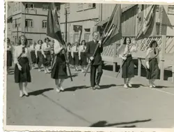 Uma fotografia em preto e branco que retrata um desfile cívico ou escolar em uma rua com construções de madeira típicas do sul do Brasil. O grupo é composto por jovens mulheres uniformizadas e um homem adulto carregando bandeiras, sugerindo um evento comemorativo de meados do século XX.