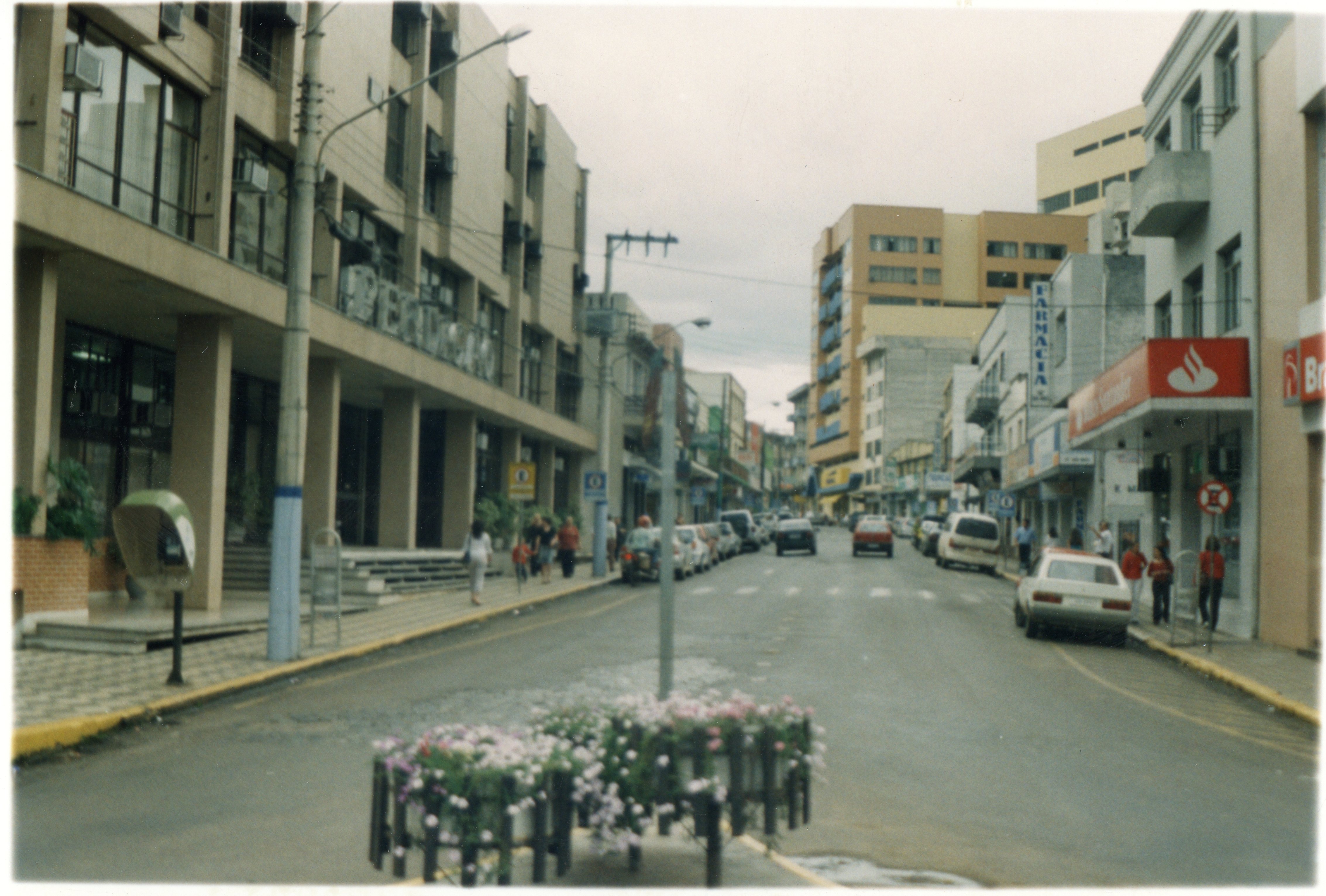 Uma vista de rua urbana em uma cidade brasileira, possivelmente Videira, caracterizada por edifícios comerciais de médio porte e tráfego de veículos. A imagem apresenta elementos típicos do final do século XX, como orelhões, sinalização de farmácia e arquitetura funcionalista.