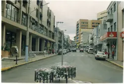 Uma vista de rua urbana em uma cidade brasileira, possivelmente Videira, caracterizada por edifícios comerciais de médio porte e tráfego de veículos. A imagem apresenta elementos típicos do final do século XX, como orelhões, sinalização de farmácia e arquitetura funcionalista.