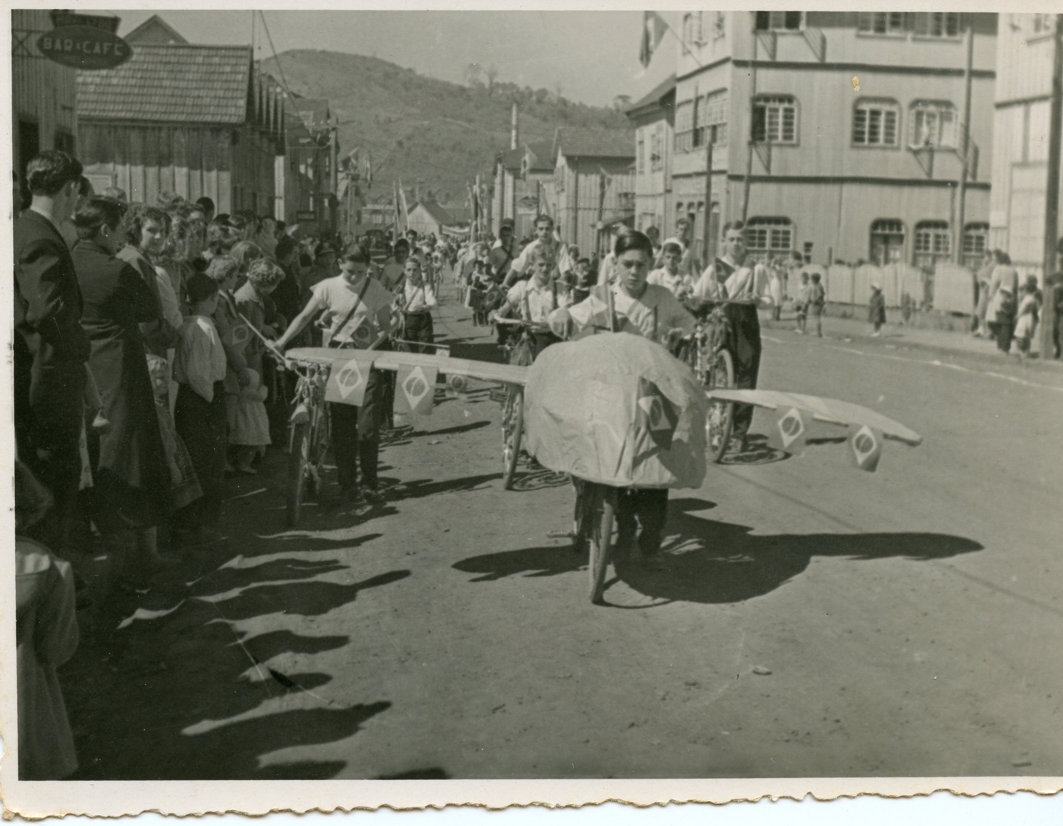 Uma fotografia em preto e branco que retrata um desfile cívico em uma rua de terra, com jovens conduzindo bicicletas decoradas como aviões e adornadas com bandeiras do Brasil. O cenário apresenta construções de madeira típicas de cidades em processo de colonização no sul do Brasil, sugerindo um evento comemorativo de meados do século XX.