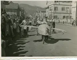 Uma fotografia em preto e branco que retrata um desfile cívico em uma rua de terra, com jovens conduzindo bicicletas decoradas como aviões e adornadas com bandeiras do Brasil. O cenário apresenta construções de madeira típicas de cidades em processo de colonização no sul do Brasil, sugerindo um evento comemorativo de meados do século XX.