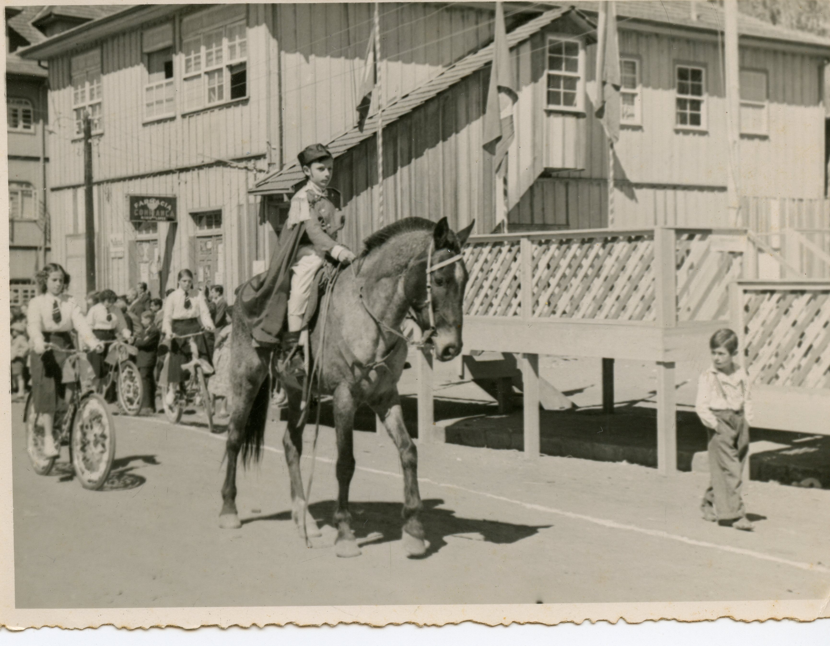 Uma fotografia em preto e branco que retrata um desfile ou evento cívico em uma pequena cidade brasileira, possivelmente na década de 1940 ou 1950. Destaca-se uma criança montada a cavalo vestindo um uniforme militar estilizado, acompanhada por jovens em bicicletas e uma arquitetura de madeira típica da colonização sulista.