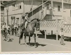 Uma fotografia em preto e branco que retrata um desfile ou evento cívico em uma pequena cidade brasileira, possivelmente na década de 1940 ou 1950. Destaca-se uma criança montada a cavalo vestindo um uniforme militar estilizado, acompanhada por jovens em bicicletas e uma arquitetura de madeira típica da colonização sulista.