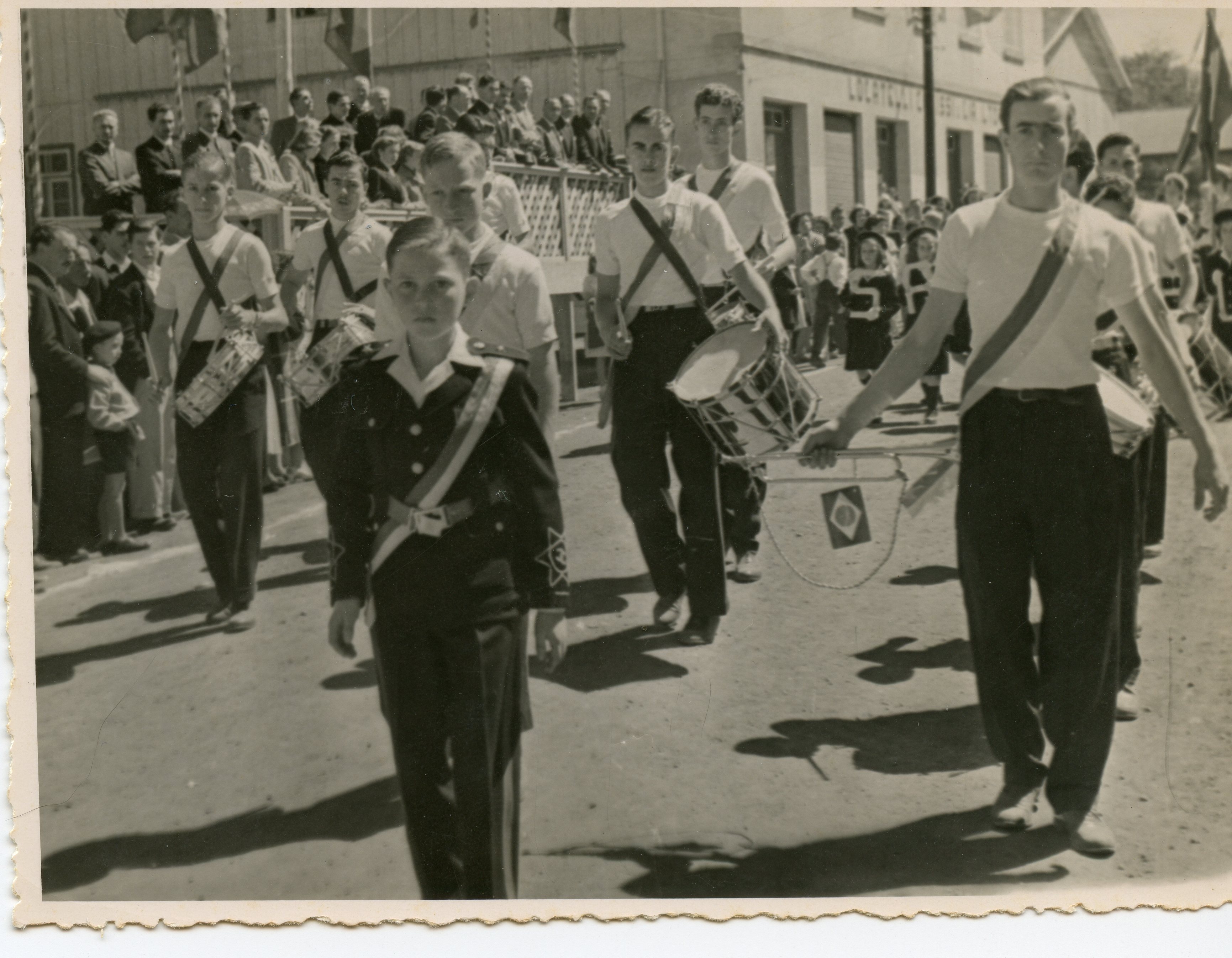 Uma fotografia em preto e branco que retrata um desfile cívico ou escolar, com jovens músicos marchando e tocando tambores. O cenário sugere uma pequena cidade brasileira, possivelmente em meados do século XX, com espectadores observando de uma arquibancada improvisada em frente a um edifício comercial.