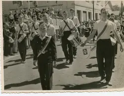 Uma fotografia em preto e branco que retrata um desfile cívico ou escolar, com jovens músicos marchando e tocando tambores. O cenário sugere uma pequena cidade brasileira, possivelmente em meados do século XX, com espectadores observando de uma arquibancada improvisada em frente a um edifício comercial.