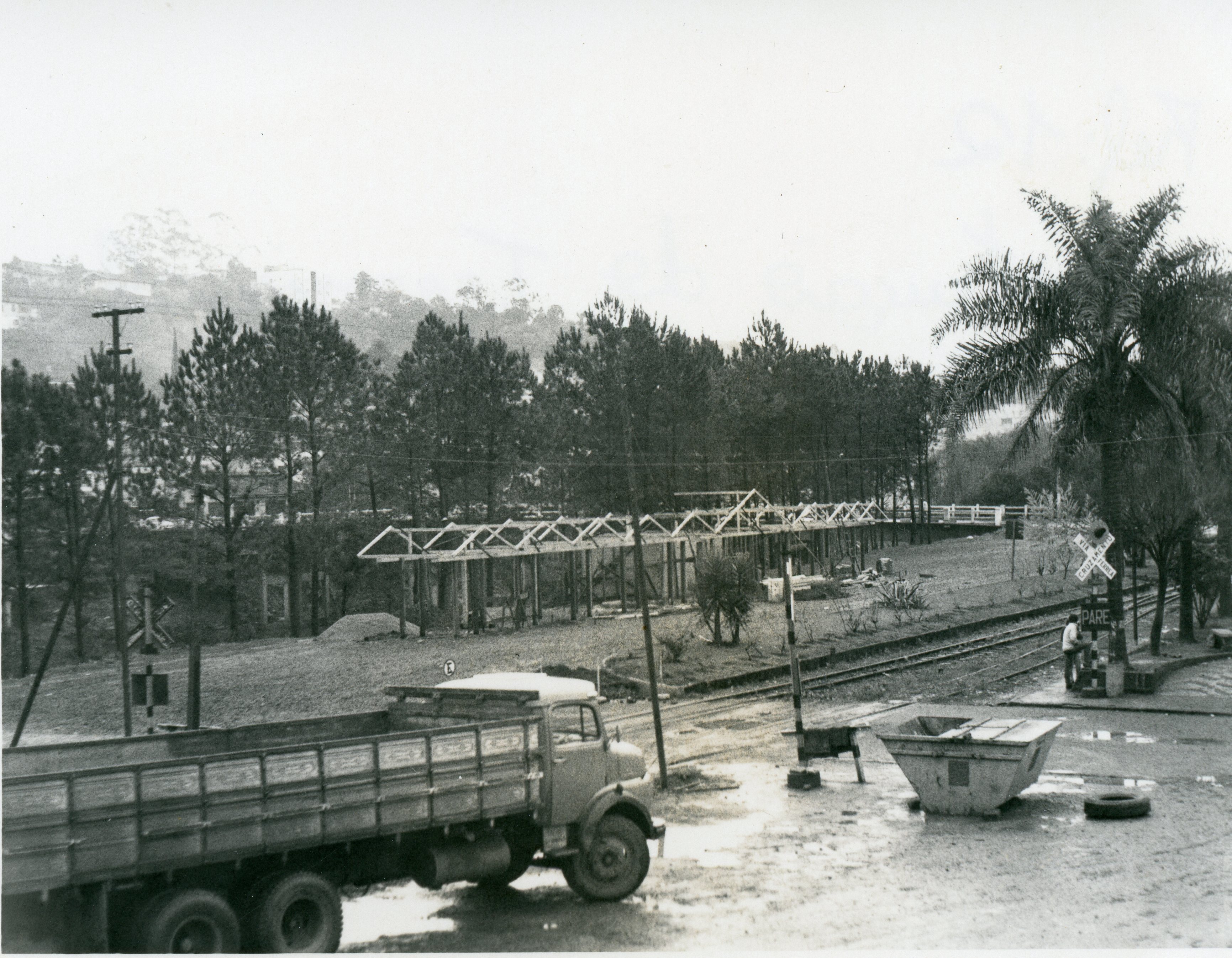 Fotografia em preto e branco mostrando uma área urbana próxima a uma linha férrea, com a estrutura de madeira de uma construção em andamento ao fundo. Em primeiro plano, observa-se um caminhão de carga estacionado em um terreno de terra batida, com uma placa de sinalização ferroviária e uma palmeira compondo a paisagem.