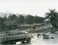 Fotografia em preto e branco mostrando uma área urbana próxima a uma linha férrea, com a estrutura de madeira de uma construção em andamento ao fundo. Em primeiro plano, observa-se um caminhão de carga estacionado em um terreno de terra batida, com uma placa de sinalização ferroviária e uma palmeira compondo a paisagem.