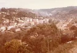 Uma vista panorâmica de uma cidade brasileira, possivelmente Videira, caracterizada por um vale cercado por colinas e uma densa ocupação urbana com edifícios de médio porte. A fotografia apresenta tons sépia típicos de registros fotográficos das décadas de 1970 ou 1980, exibindo uma paisagem em desenvolvimento com vegetação preservada nas encostas.