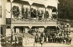 Fotografia histórica de um evento social em frente ao Clube União, um edifício de madeira com arquitetura típica de colonização europeia no sul do Brasil. A cena mostra uma grande multidão reunida, com pessoas no térreo e na sacada superior, decorada com festões e folhagens, sugerindo uma celebração comunitária ou festividade local da primeira metade do século XX.