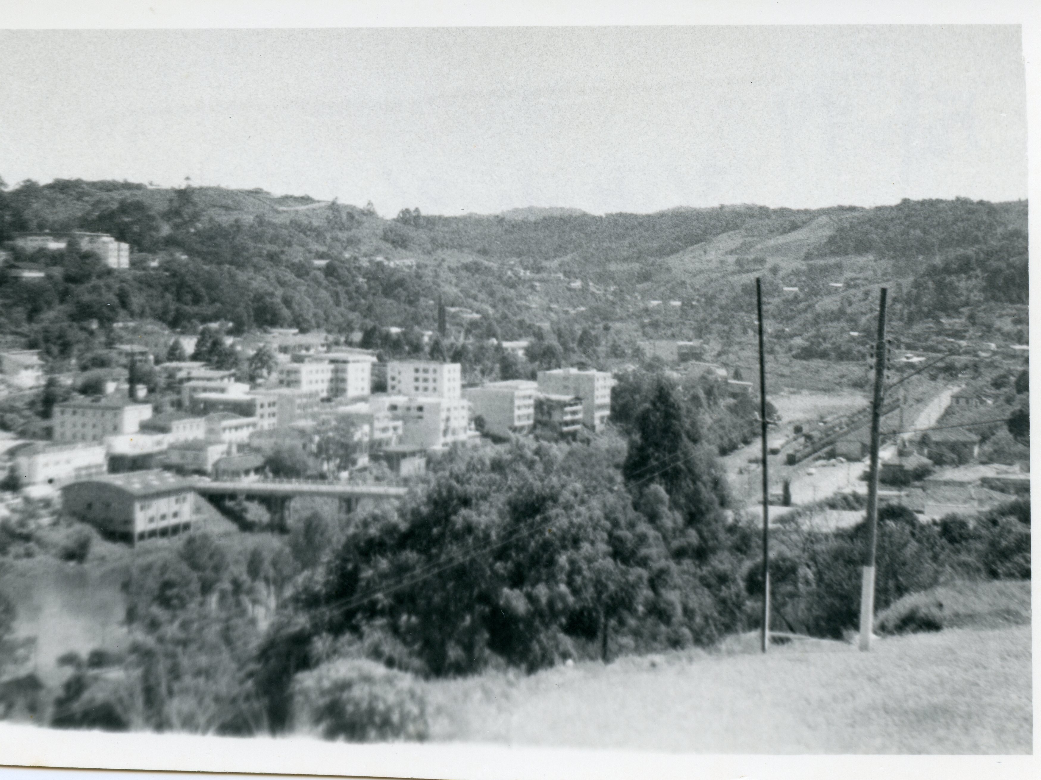 Uma fotografia em preto e branco que apresenta uma vista panorâmica de uma pequena cidade brasileira, possivelmente Videira, situada em um vale cercado por colinas arborizadas. A imagem exibe um centro urbano em desenvolvimento com edifícios de arquitetura funcionalista, uma ponte sobre um curso d'água e postes de energia, sugerindo um período de expansão urbana entre as décadas de 1960 e 1970.