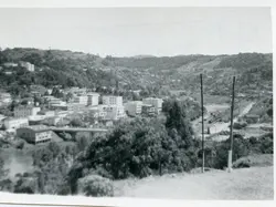 Uma fotografia em preto e branco que apresenta uma vista panorâmica de uma pequena cidade brasileira, possivelmente Videira, situada em um vale cercado por colinas arborizadas. A imagem exibe um centro urbano em desenvolvimento com edifícios de arquitetura funcionalista, uma ponte sobre um curso d'água e postes de energia, sugerindo um período de expansão urbana entre as décadas de 1960 e 1970.