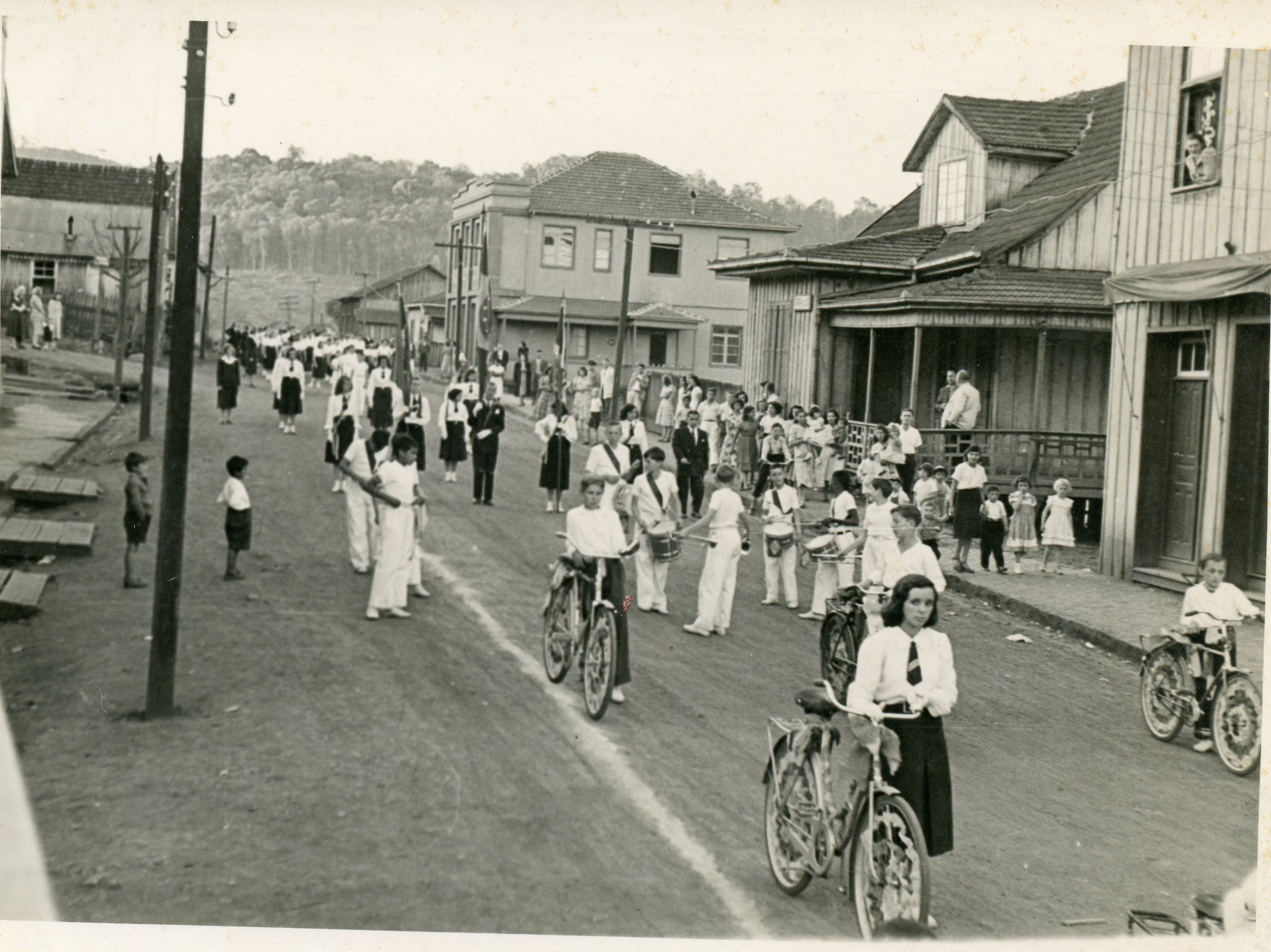 Uma fotografia em preto e branco que retrata um desfile cívico ou procissão em uma rua de terra de uma pequena cidade brasileira, possivelmente no sul do país devido à arquitetura de madeira. Observam-se jovens uniformizados, alguns com instrumentos musicais e bicicletas, em um cenário que remete à primeira metade do século XX.
