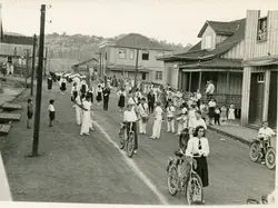 Uma fotografia em preto e branco que retrata um desfile cívico ou procissão em uma rua de terra de uma pequena cidade brasileira, possivelmente no sul do país devido à arquitetura de madeira. Observam-se jovens uniformizados, alguns com instrumentos musicais e bicicletas, em um cenário que remete à primeira metade do século XX.