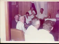 Uma fotografia colorida de época, provavelmente das décadas de 1970 ou 1980, retratando um grupo de homens reunidos em um ambiente interno com revestimento de madeira. O cenário sugere uma confraternização social ou reunião informal, com a presença de bebidas sobre uma mesa, refletindo um costume comum em clubes ou residências da época.