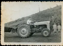 Fotografia em preto e branco retratando um jovem operando um trator Ford com implemento agrícola em uma área rural. Ao lado, duas crianças observam a cena, sugerindo um contexto de vida no campo, possivelmente na metade do século XX.