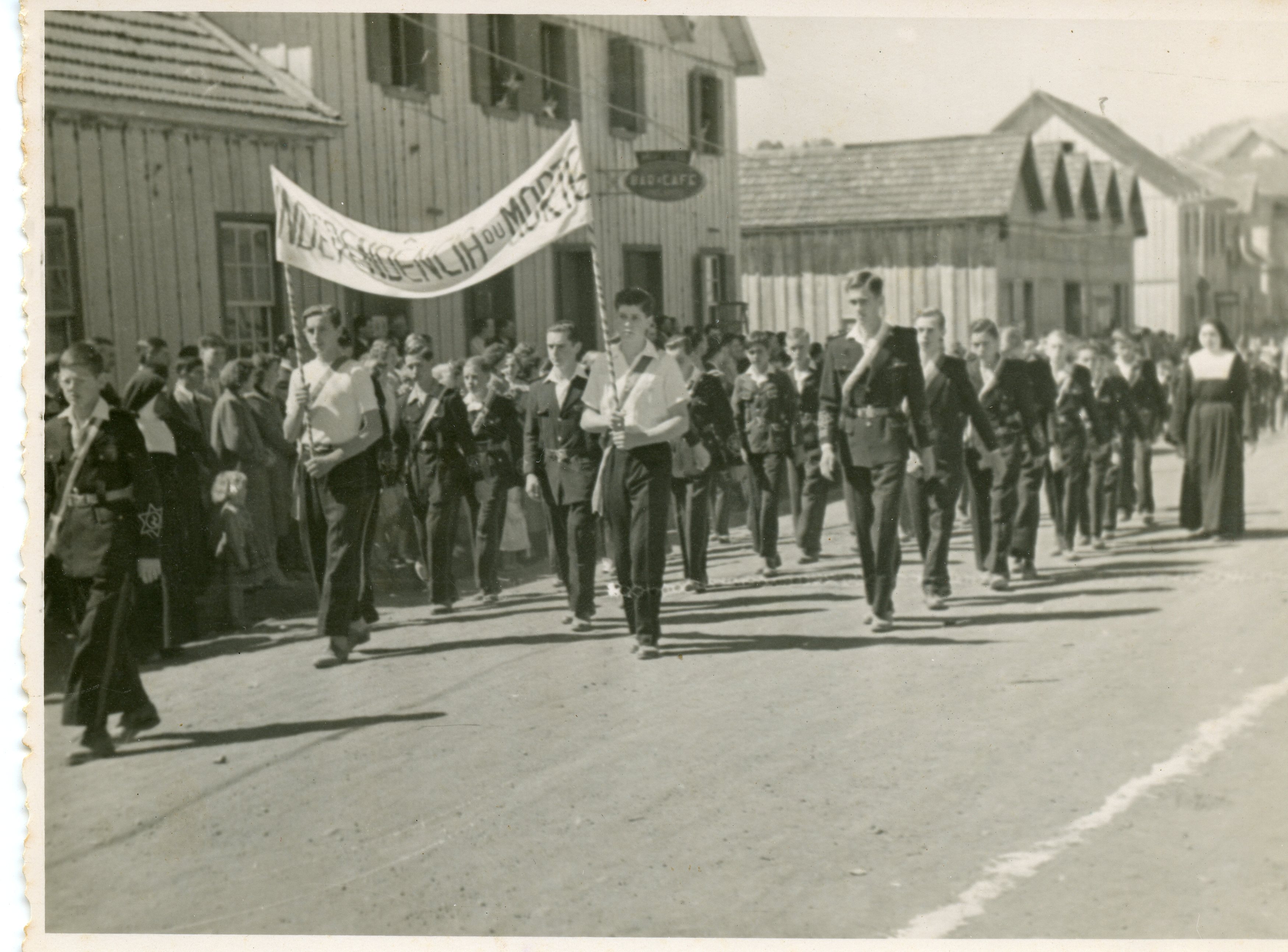 Fotografia em preto e branco de um desfile cívico em uma pequena cidade brasileira, possivelmente em meados do século XX. Jovens estudantes marcham em uma rua de terra, carregando uma faixa com a inscrição 'Independência ou Morte', com casas de madeira típicas da colonização ao fundo.