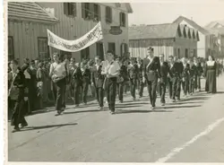 Fotografia em preto e branco de um desfile cívico em uma pequena cidade brasileira, possivelmente em meados do século XX. Jovens estudantes marcham em uma rua de terra, carregando uma faixa com a inscrição 'Independência ou Morte', com casas de madeira típicas da colonização ao fundo.