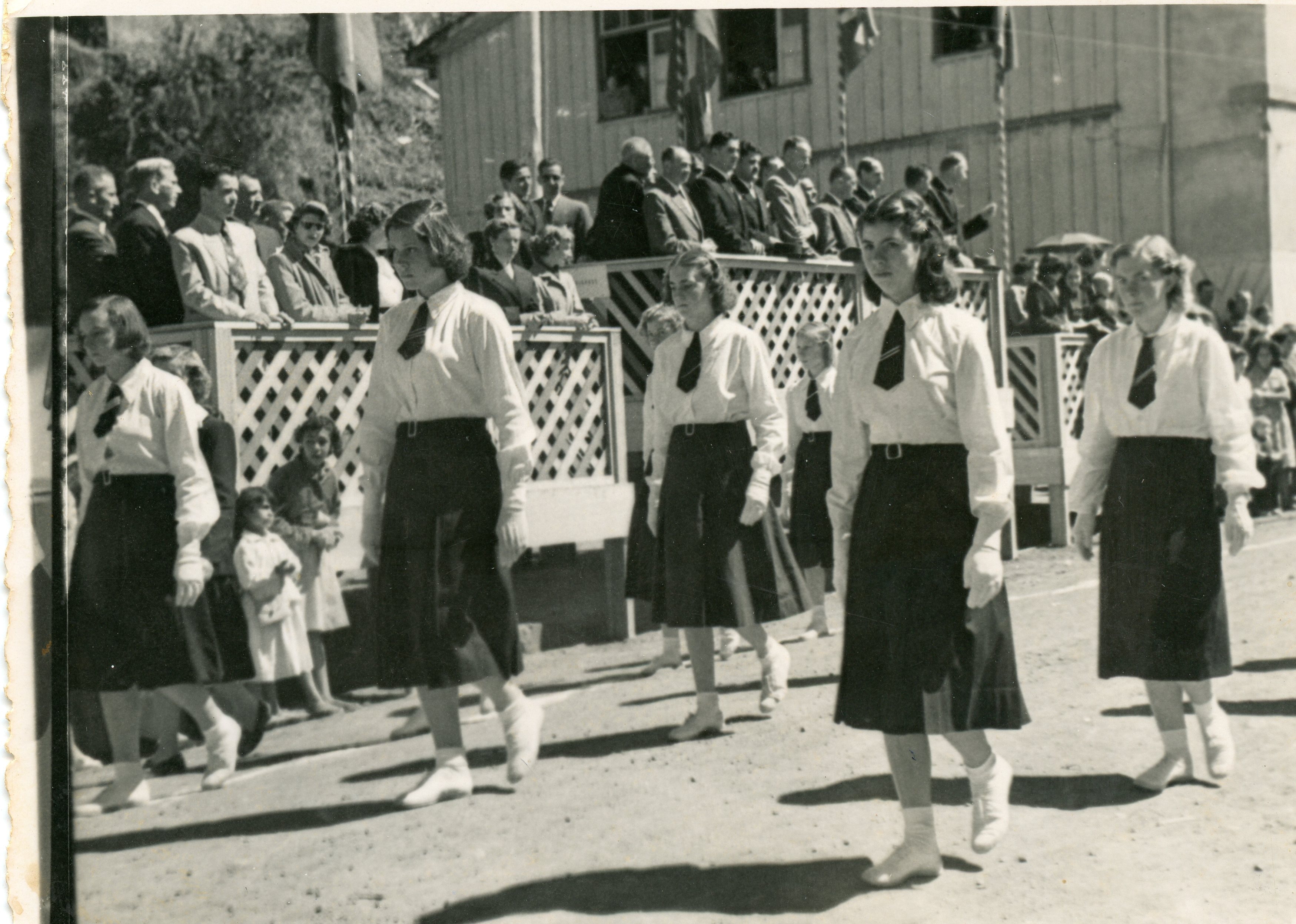 Uma fotografia em preto e branco que retrata um desfile cívico ou escolar, com jovens estudantes marchando uniformizadas com camisas brancas, saias escuras e gravatas. Ao fundo, observa-se uma estrutura de madeira com arquibancadas onde adultos assistem ao evento, sugerindo um contexto de celebração comunitária ou escolar em uma pequena cidade brasileira de meados do século XX.