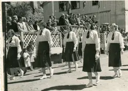 Uma fotografia em preto e branco que retrata um desfile cívico ou escolar, com jovens estudantes marchando uniformizadas com camisas brancas, saias escuras e gravatas. Ao fundo, observa-se uma estrutura de madeira com arquibancadas onde adultos assistem ao evento, sugerindo um contexto de celebração comunitária ou escolar em uma pequena cidade brasileira de meados do século XX.