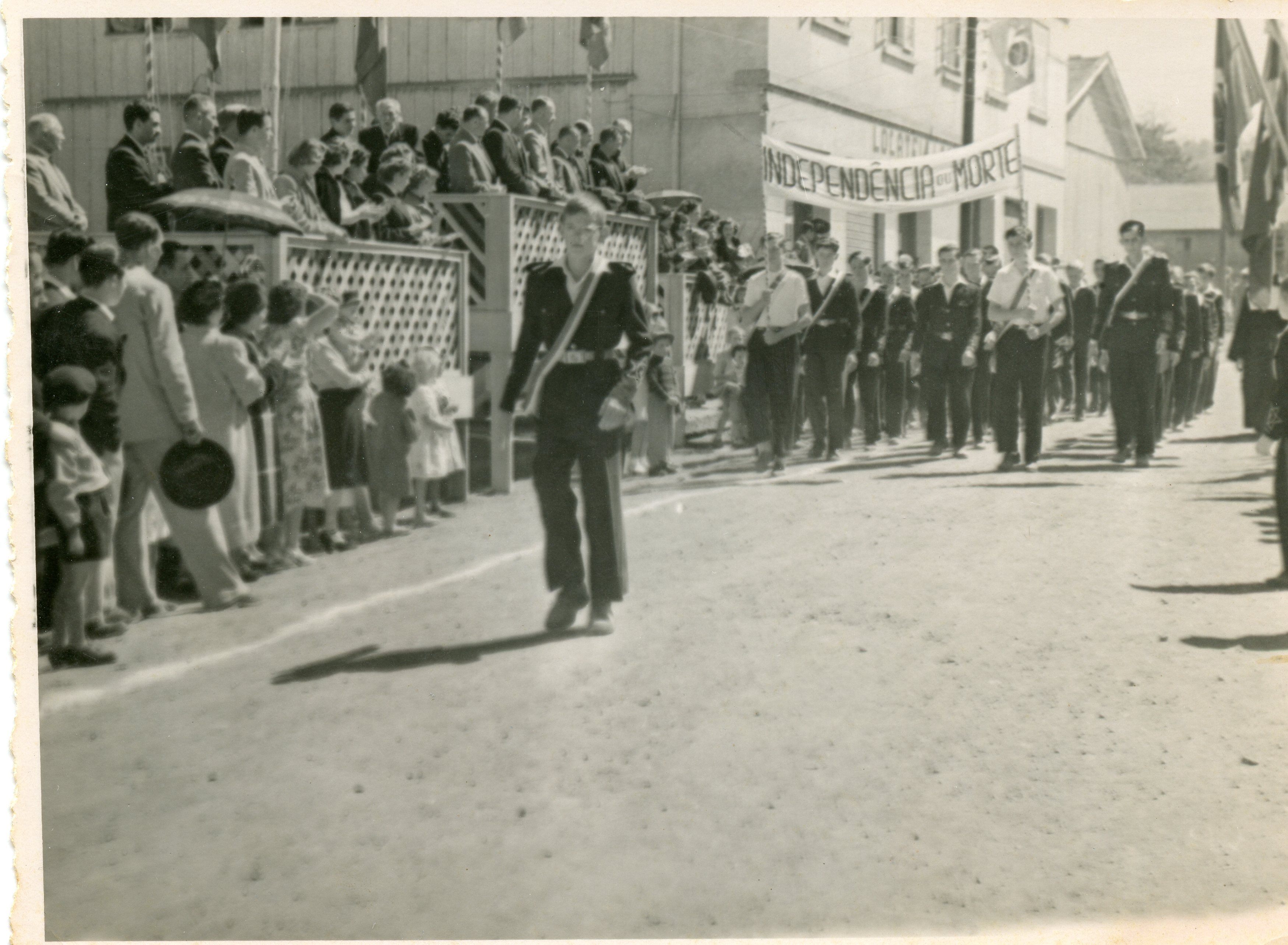 Fotografia em preto e branco de um desfile cívico, provavelmente em comemoração ao 7 de setembro, em uma pequena cidade brasileira. Observa-se um grupo de jovens marchando com uma faixa onde se lê 'Independência ou Morte', enquanto espectadores acompanham o evento atrás de uma cerca de madeira.