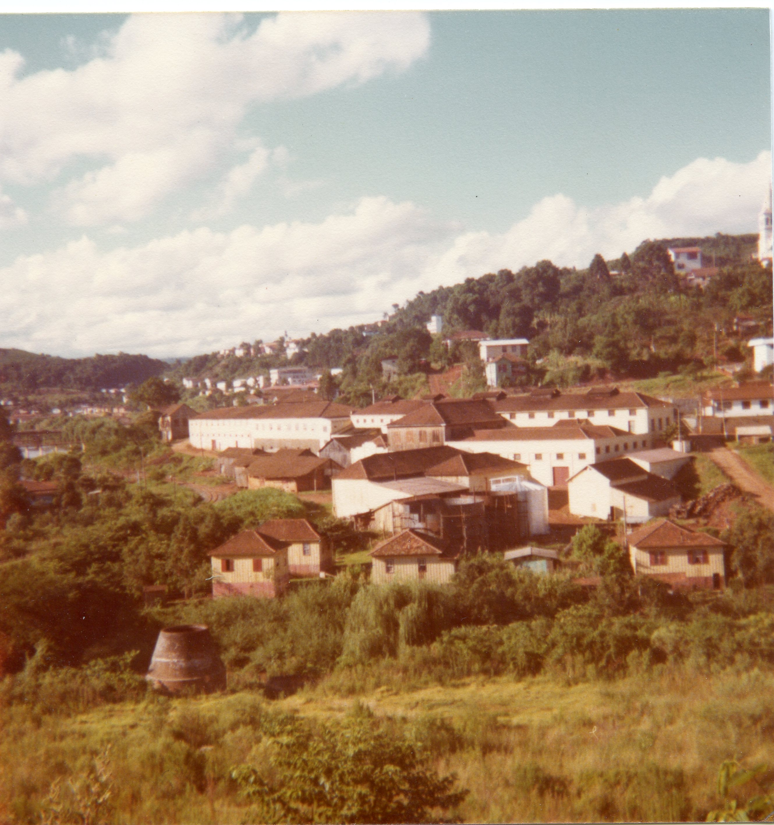 Uma vista panorâmica de uma pequena cidade brasileira, possivelmente Videira, destacando um complexo industrial ou armazéns cercados por vegetação e casas simples. A fotografia, com tons levemente desbotados típicos da década de 1970 ou 1980, mostra uma paisagem urbana em desenvolvimento com colinas ao fundo.