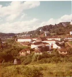 Uma vista panorâmica de uma pequena cidade brasileira, possivelmente Videira, destacando um complexo industrial ou armazéns cercados por vegetação e casas simples. A fotografia, com tons levemente desbotados típicos da década de 1970 ou 1980, mostra uma paisagem urbana em desenvolvimento com colinas ao fundo.