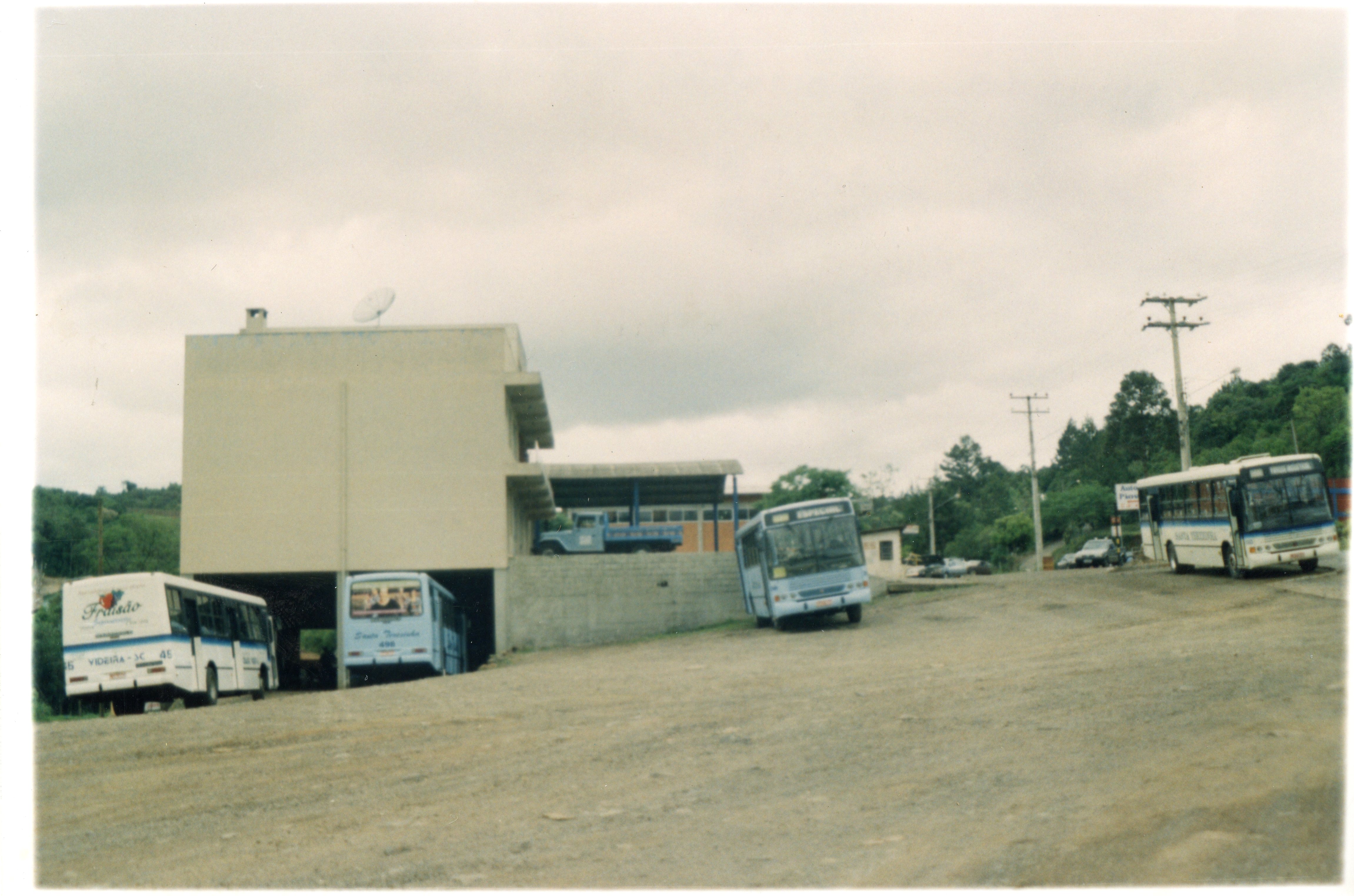 Uma fotografia de um terminal rodoviário ou pátio de manobras em Videira, Santa Catarina, apresentando vários ônibus estacionados em um terreno de terra batida. O cenário inclui um prédio de alvenaria com arquitetura funcionalista típica de meados da década de 1990, refletindo o transporte público regional da época.