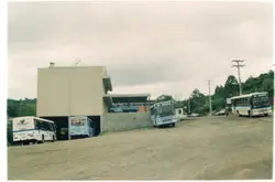 Uma fotografia de um terminal rodoviário ou pátio de manobras em Videira, Santa Catarina, apresentando vários ônibus estacionados em um terreno de terra batida. O cenário inclui um prédio de alvenaria com arquitetura funcionalista típica de meados da década de 1990, refletindo o transporte público regional da época.