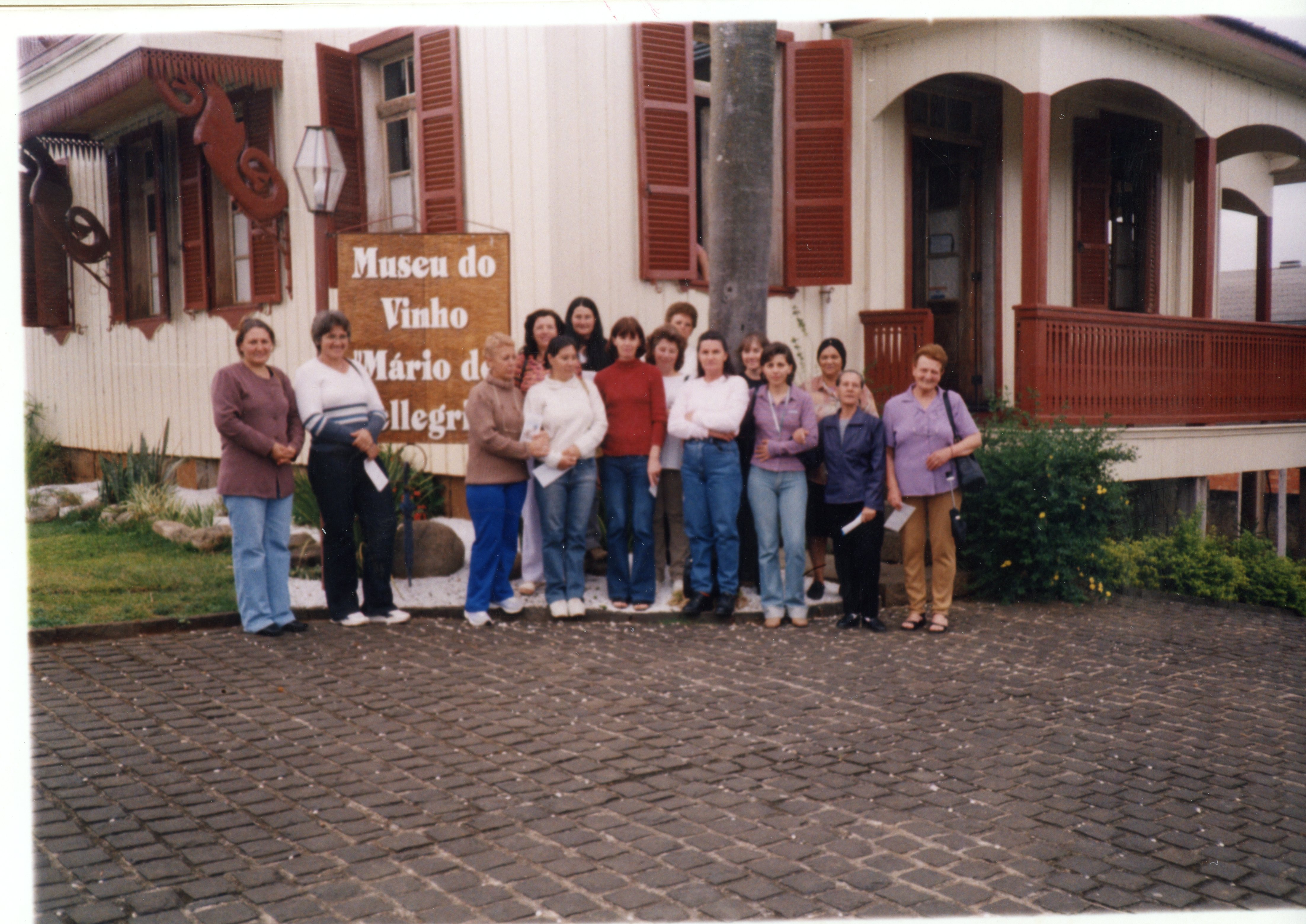 Uma fotografia colorida mostrando um grupo de mulheres posando em frente ao Museu do Vinho Mário de Pellegrini, em Videira, Santa Catarina. A arquitetura de madeira com varanda e detalhes em vermelho sugere uma construção histórica preservada, enquanto as vestimentas das pessoas indicam um período entre o final da década de 1990 e início dos anos 2000.