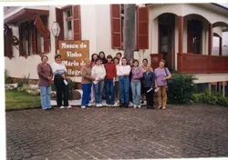 Uma fotografia colorida mostrando um grupo de mulheres posando em frente ao Museu do Vinho Mário de Pellegrini, em Videira, Santa Catarina. A arquitetura de madeira com varanda e detalhes em vermelho sugere uma construção histórica preservada, enquanto as vestimentas das pessoas indicam um período entre o final da década de 1990 e início dos anos 2000.