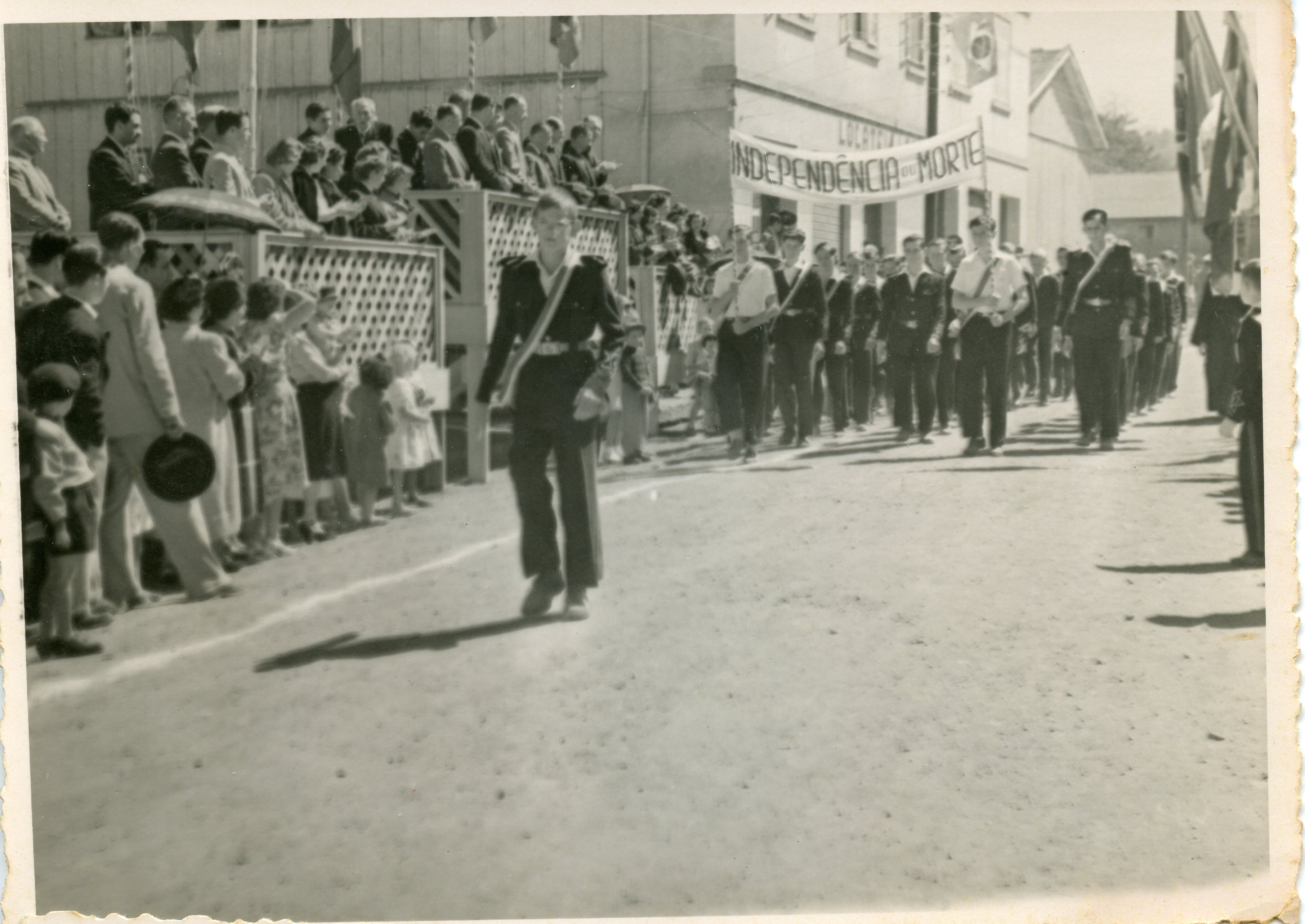 Uma fotografia em preto e branco que registra um desfile cívico, provavelmente em comemoração ao 7 de setembro, em uma pequena cidade brasileira. O grupo de jovens marcha em uma rua de terra, carregando uma faixa com os dizeres 'Independência ou Morte', enquanto espectadores observam de uma arquibancada improvisada de madeira.