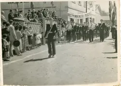 Uma fotografia em preto e branco que registra um desfile cívico, provavelmente em comemoração ao 7 de setembro, em uma pequena cidade brasileira. O grupo de jovens marcha em uma rua de terra, carregando uma faixa com os dizeres 'Independência ou Morte', enquanto espectadores observam de uma arquibancada improvisada de madeira.