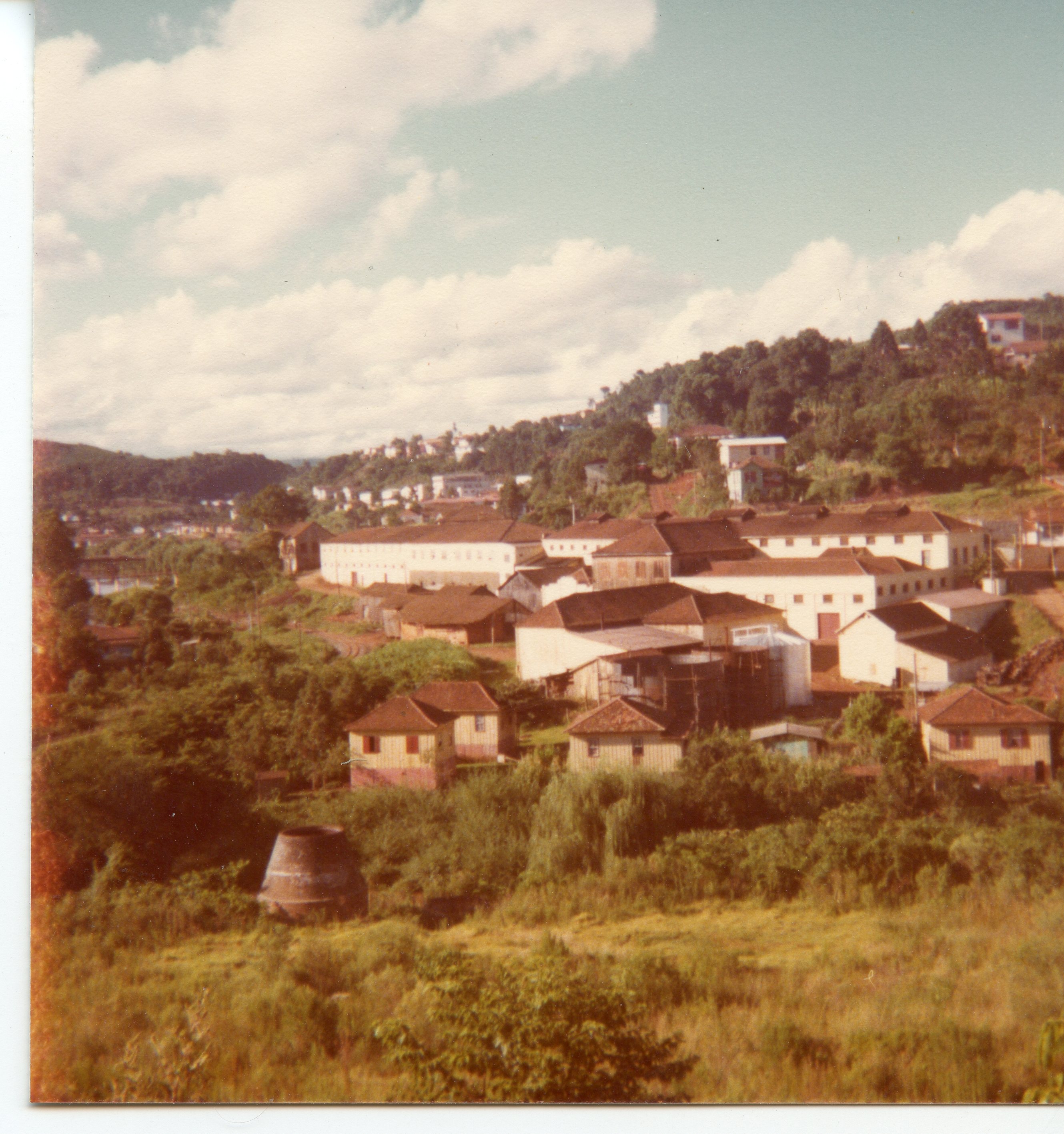 Uma vista panorâmica de uma pequena cidade brasileira, possivelmente Videira, caracterizada por um complexo industrial ou armazéns de grande porte cercados por casas de madeira e vegetação densa. A fotografia, com tons envelhecidos típicos da década de 1970, mostra uma paisagem urbana em desenvolvimento integrada à topografia montanhosa.