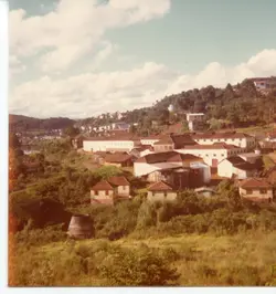 Uma vista panorâmica de uma pequena cidade brasileira, possivelmente Videira, caracterizada por um complexo industrial ou armazéns de grande porte cercados por casas de madeira e vegetação densa. A fotografia, com tons envelhecidos típicos da década de 1970, mostra uma paisagem urbana em desenvolvimento integrada à topografia montanhosa.