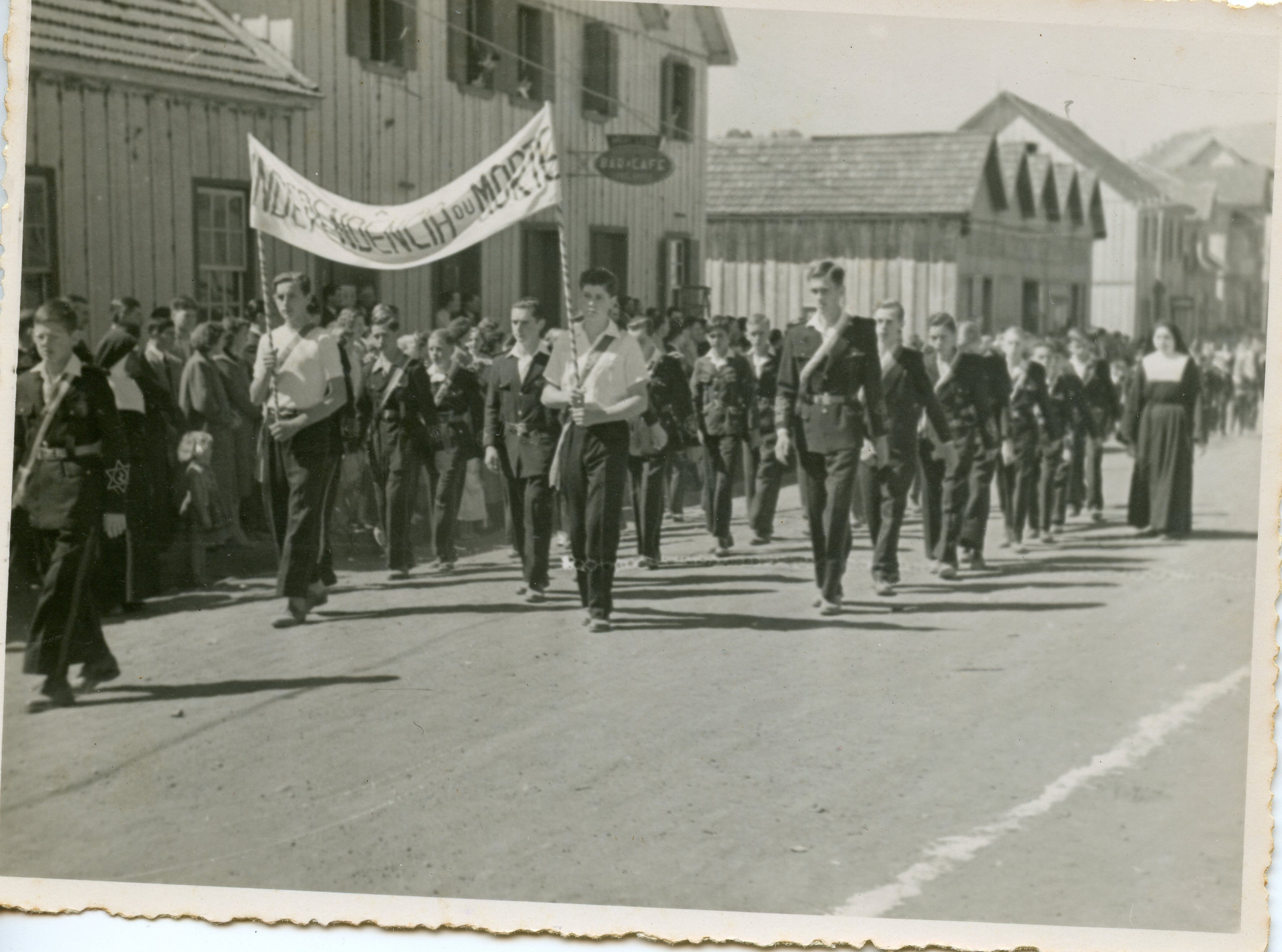 Fotografia em preto e branco de um desfile cívico, provavelmente em uma cidade pequena do interior do Brasil, com jovens marchando e carregando uma faixa com os dizeres 'Independência ou Morte'. A arquitetura de madeira das casas ao fundo sugere um período de colonização ou meados do século XX, com a presença de uma freira e estudantes uniformizados.