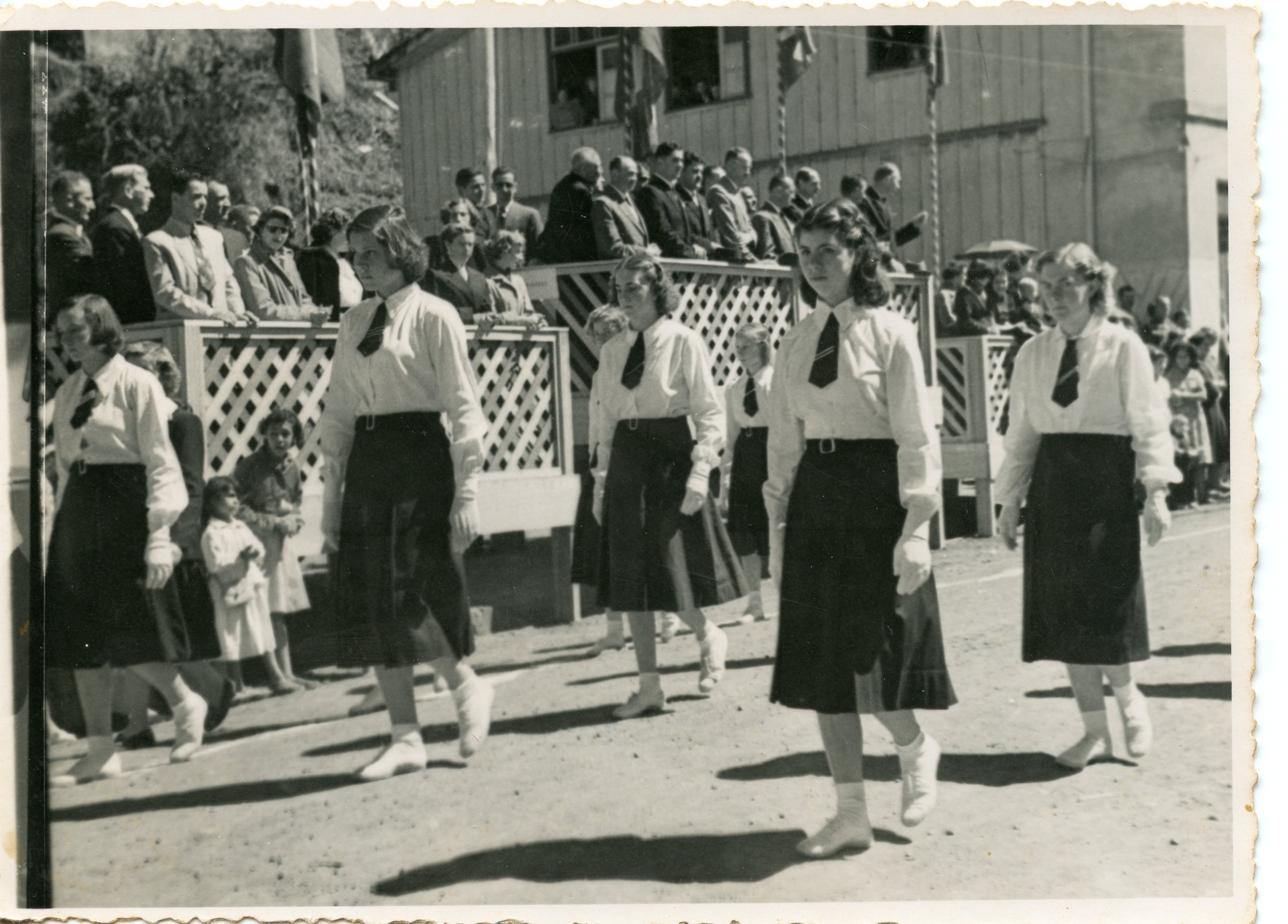Uma fotografia em preto e branco que retrata um desfile cívico ou escolar, com jovens mulheres vestindo uniformes compostos por camisas brancas, saias escuras e gravatas. Ao fundo, observa-se uma estrutura de madeira com treliças e uma multidão de espectadores, sugerindo um evento comunitário em uma pequena cidade brasileira de meados do século XX.
