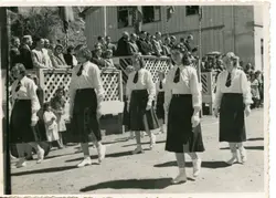 Uma fotografia em preto e branco que retrata um desfile cívico ou escolar, com jovens mulheres vestindo uniformes compostos por camisas brancas, saias escuras e gravatas. Ao fundo, observa-se uma estrutura de madeira com treliças e uma multidão de espectadores, sugerindo um evento comunitário em uma pequena cidade brasileira de meados do século XX.