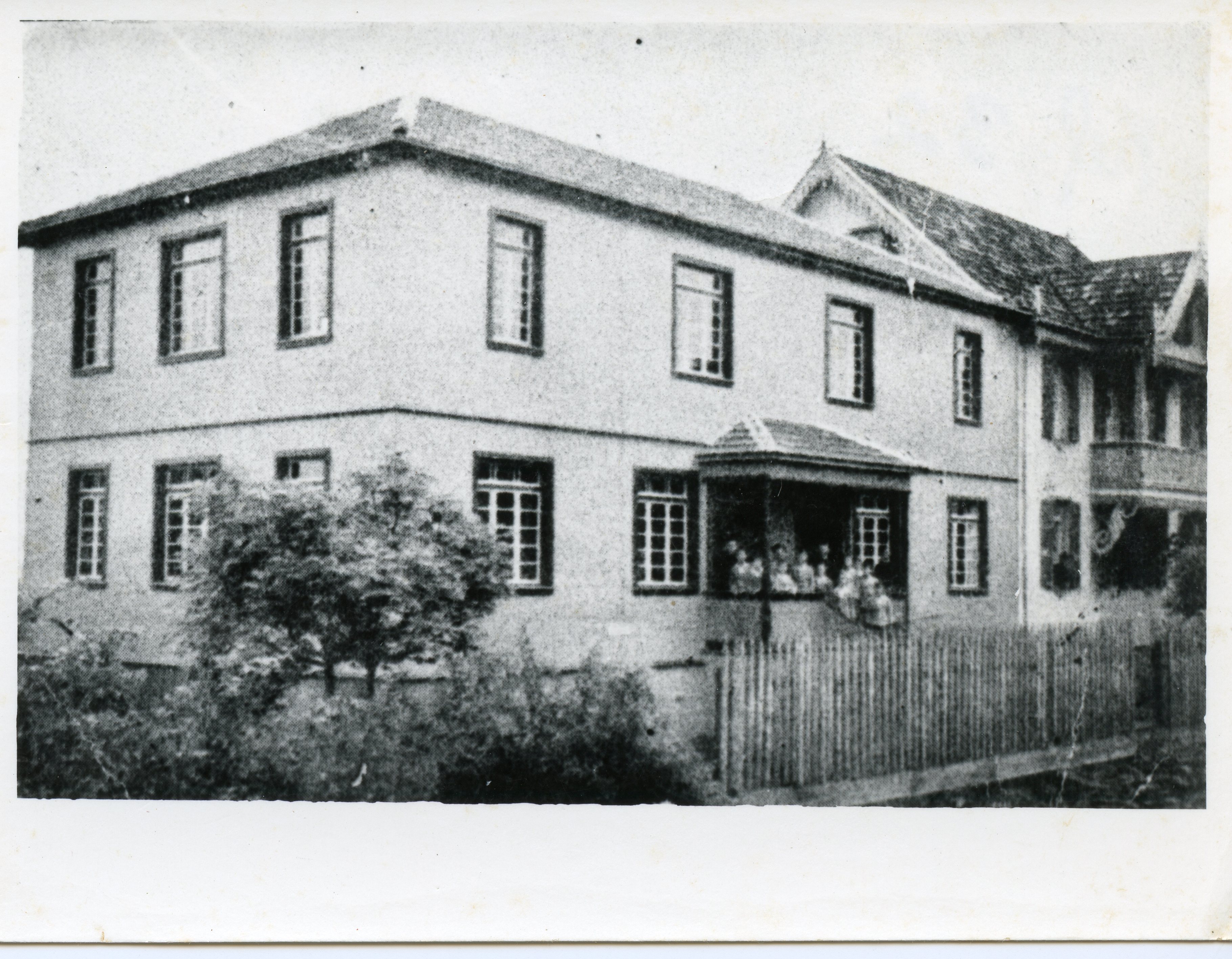 Fotografia em preto e branco de um casarão de dois andares com arquitetura típica de colonização europeia, possivelmente no sul do Brasil. Observa-se um grupo de pessoas sentado na varanda frontal, protegida por uma cerca de madeira, sugerindo um ambiente residencial ou institucional do início do século XX.