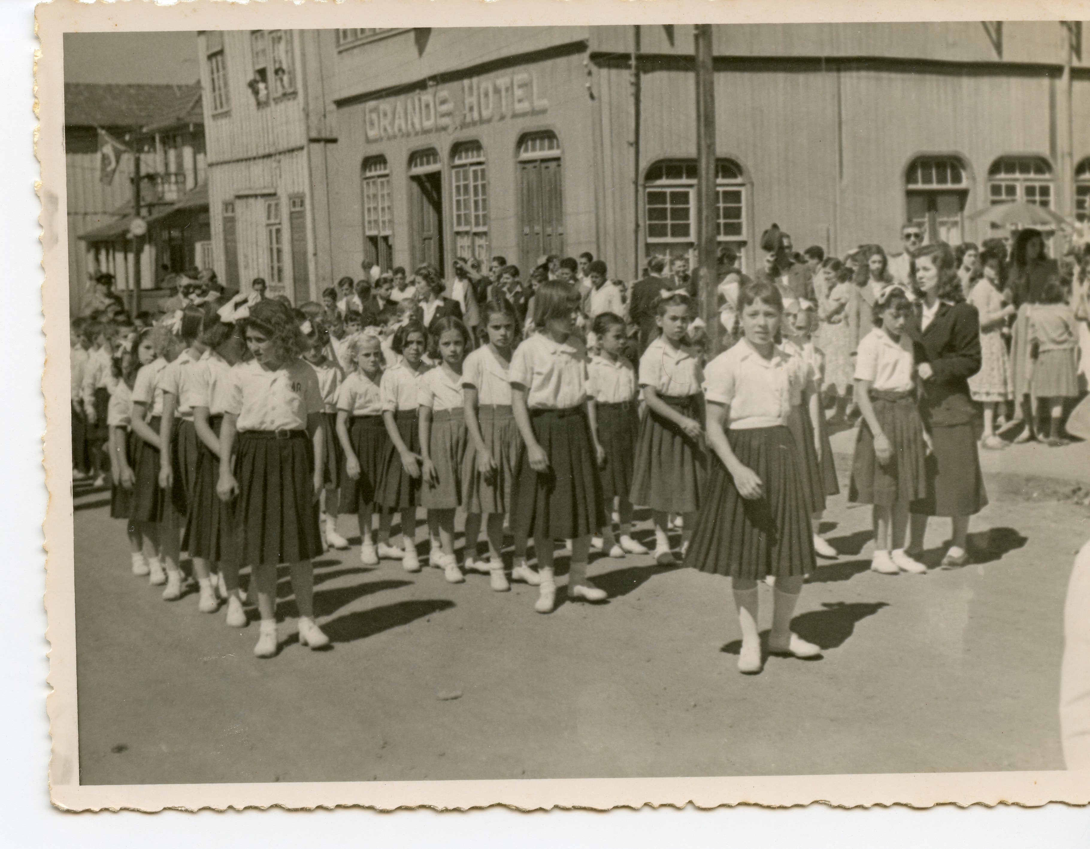 Fotografia em preto e branco retratando um desfile cívico ou escolar em uma pequena cidade brasileira, com um grupo de meninas uniformizadas em primeiro plano. Ao fundo, destaca-se uma construção de madeira com a inscrição 'GRANDE HOTEL', sugerindo um contexto de desenvolvimento urbano no interior do Brasil, provavelmente entre as décadas de 1940 e 1950.