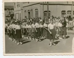 Fotografia em preto e branco retratando um desfile cívico ou escolar em uma pequena cidade brasileira, com um grupo de meninas uniformizadas em primeiro plano. Ao fundo, destaca-se uma construção de madeira com a inscrição 'GRANDE HOTEL', sugerindo um contexto de desenvolvimento urbano no interior do Brasil, provavelmente entre as décadas de 1940 e 1950.