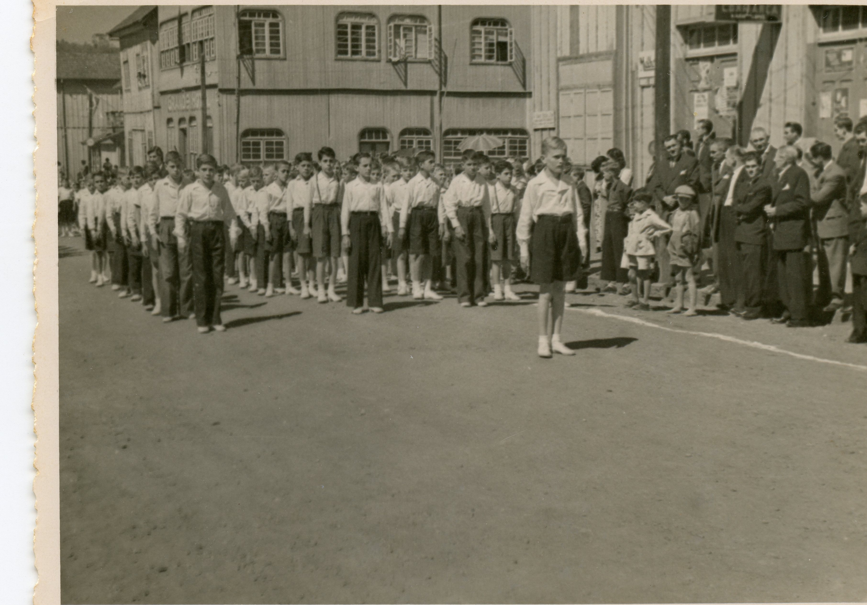 Uma fotografia em preto e branco que retrata um desfile ou evento cívico com um grupo de meninos uniformizados marchando em uma rua de terra. Ao fundo, observa-se uma arquitetura de madeira típica de cidades colonizadas por imigrantes europeus no sul do Brasil, com espectadores adultos acompanhando a cena.