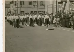 Uma fotografia em preto e branco que retrata um desfile ou evento cívico com um grupo de meninos uniformizados marchando em uma rua de terra. Ao fundo, observa-se uma arquitetura de madeira típica de cidades colonizadas por imigrantes europeus no sul do Brasil, com espectadores adultos acompanhando a cena.
