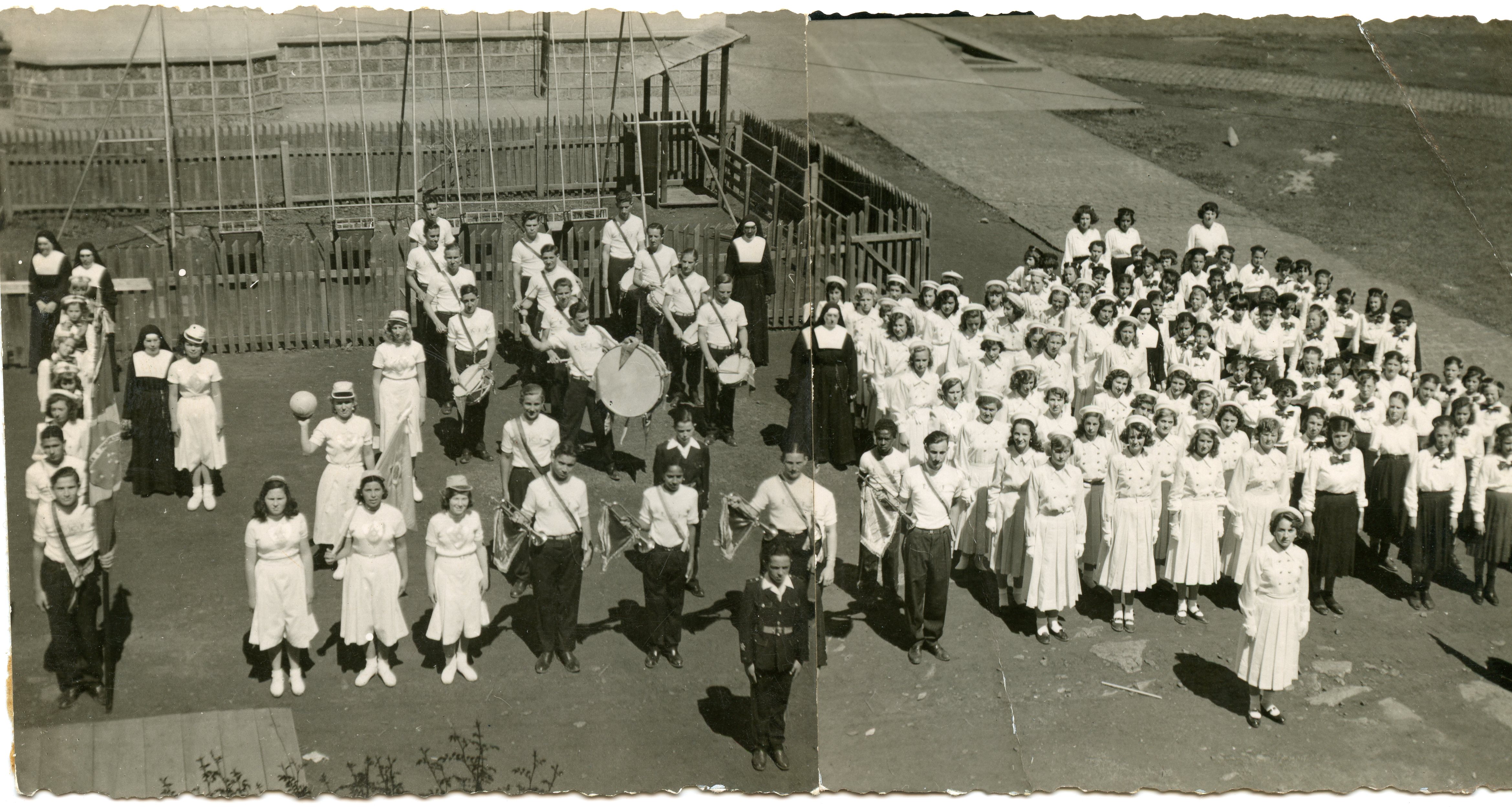 Uma fotografia em preto e branco que retrata um grande grupo de estudantes, possivelmente de um colégio católico, organizados em formação para um desfile ou evento cívico. O grupo inclui uma banda marcial com instrumentos de percussão e sopro, acompanhados por freiras, em um pátio escolar com um parquinho ao fundo, sugerindo uma cena educacional brasileira de meados do século XX.