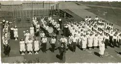 Uma fotografia em preto e branco que retrata um grande grupo de estudantes, possivelmente de um colégio católico, organizados em formação para um desfile ou evento cívico. O grupo inclui uma banda marcial com instrumentos de percussão e sopro, acompanhados por freiras, em um pátio escolar com um parquinho ao fundo, sugerindo uma cena educacional brasileira de meados do século XX.