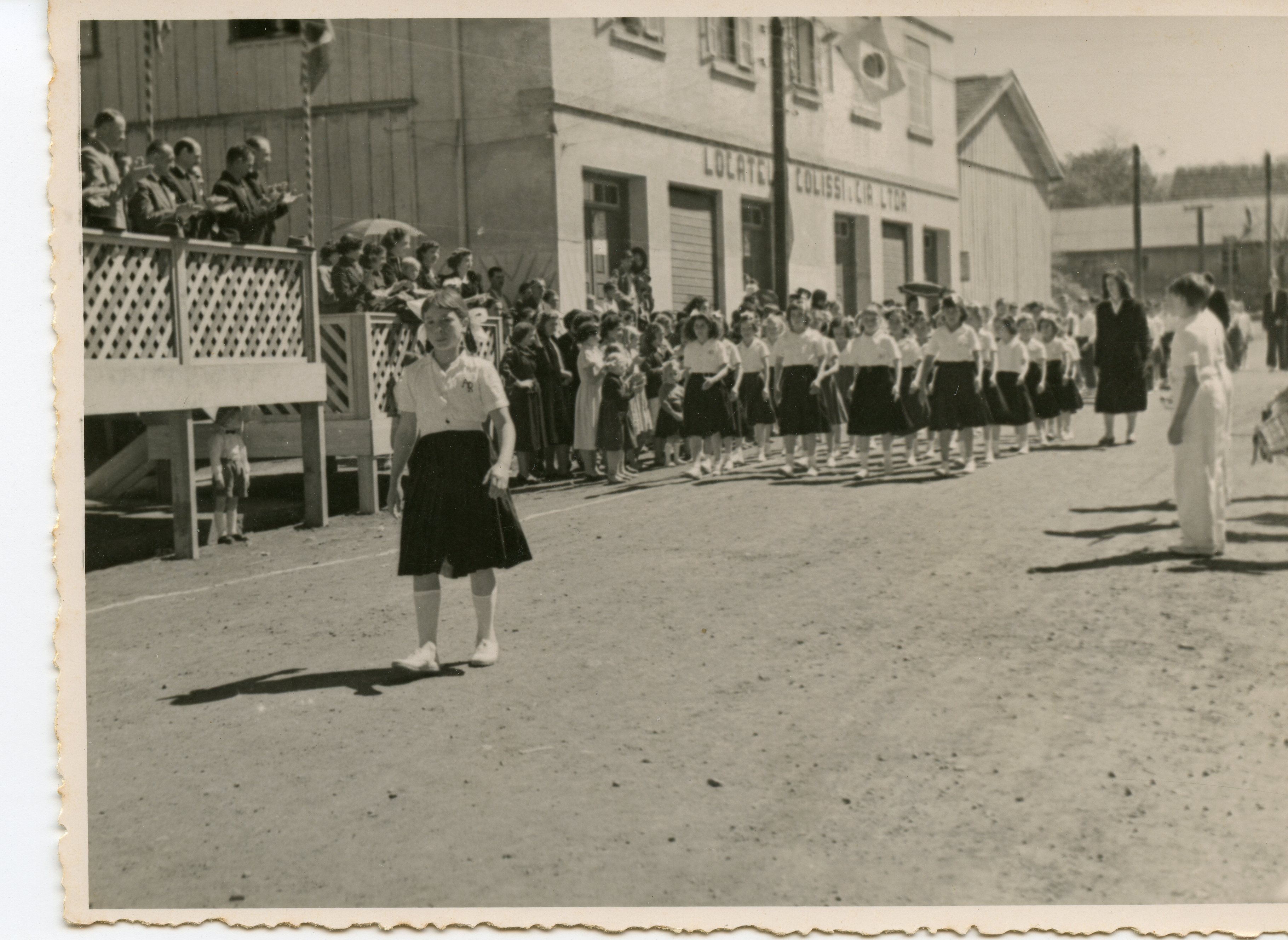 Fotografia em preto e branco de um desfile cívico ou escolar em uma rua de terra, possivelmente em uma cidade do interior do Brasil como Videira. Estudantes uniformizadas marcham em fila, enquanto espectadores observam de uma estrutura de madeira elevada e das calçadas, sugerindo um evento comunitário de meados do século XX.