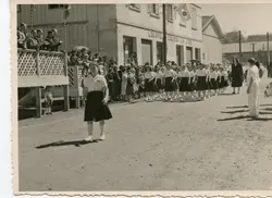 Fotografia em preto e branco de um desfile cívico ou escolar em uma rua de terra, possivelmente em uma cidade do interior do Brasil como Videira. Estudantes uniformizadas marcham em fila, enquanto espectadores observam de uma estrutura de madeira elevada e das calçadas, sugerindo um evento comunitário de meados do século XX.