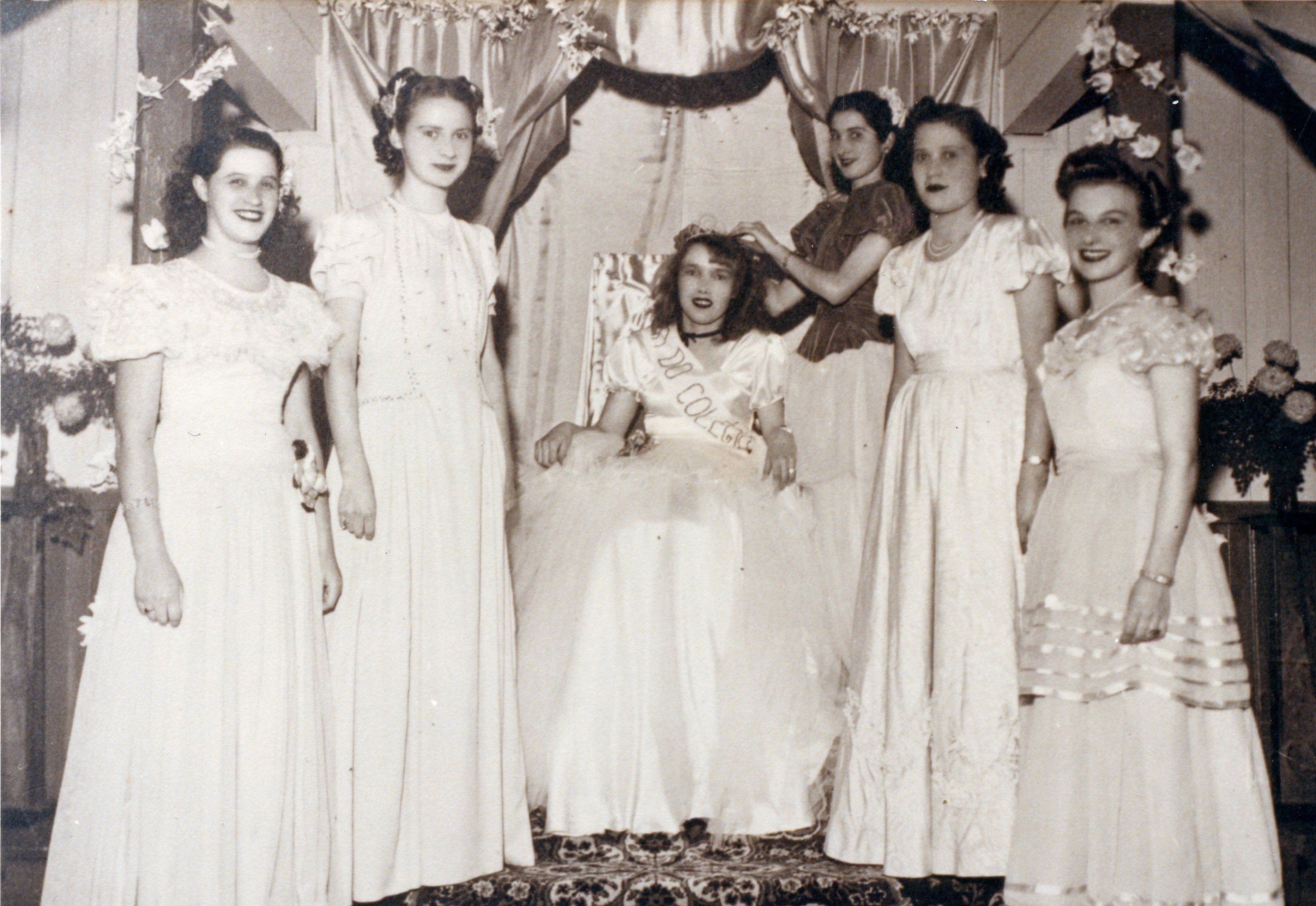 Fotografia em preto e branco retratando um grupo de jovens mulheres em um evento social, possivelmente uma coroação de rainha de colégio, dado a faixa na jovem sentada. O cenário sugere um ambiente interno decorado com cortinas e flores, típico de festividades escolares ou sociais de meados do século XX.