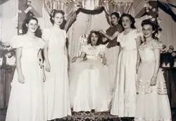 Fotografia em preto e branco retratando um grupo de jovens mulheres em um evento social, possivelmente uma coroação de rainha de colégio, dado a faixa na jovem sentada. O cenário sugere um ambiente interno decorado com cortinas e flores, típico de festividades escolares ou sociais de meados do século XX.